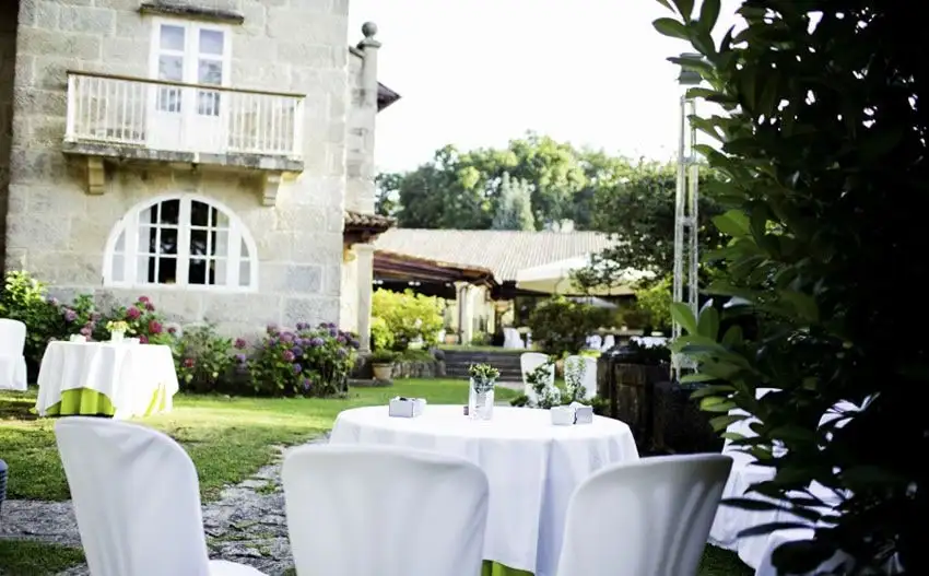 La fachada de piedra de la casona con su balcón y su ventana en arco, sobre un jardín de hortensias y mesas con mantel blanco