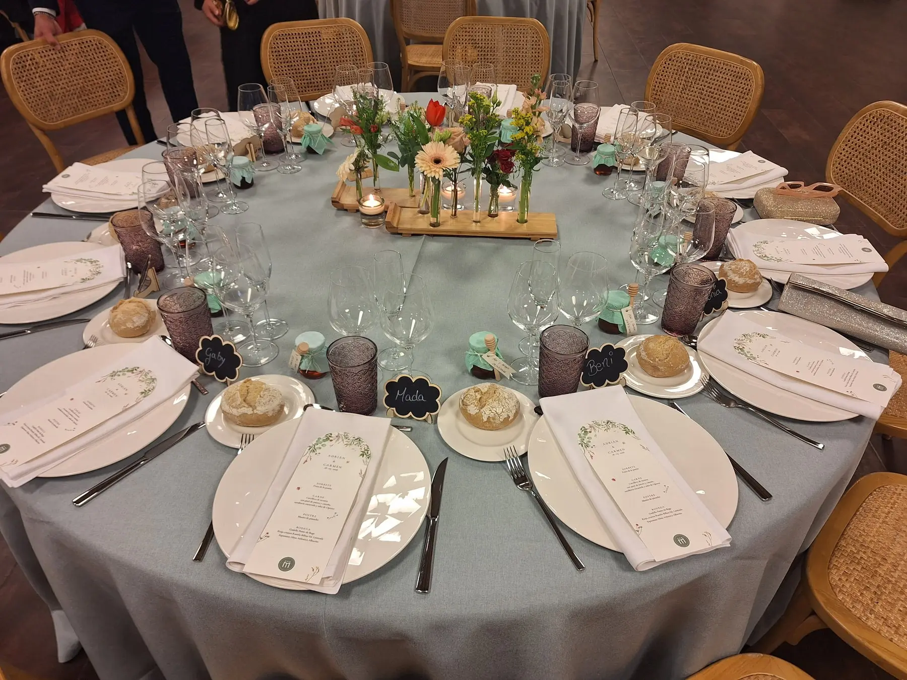 Una mesa redonda de banquete con mantel verde agua, jarroncitos de flores de colores, minutas y sillas de rejilla