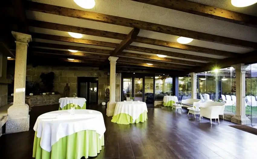 Un porche de columnas de granito y vigas de madera con mesas de mantel blanco y verde, abierto con paredes de cristal al jardín