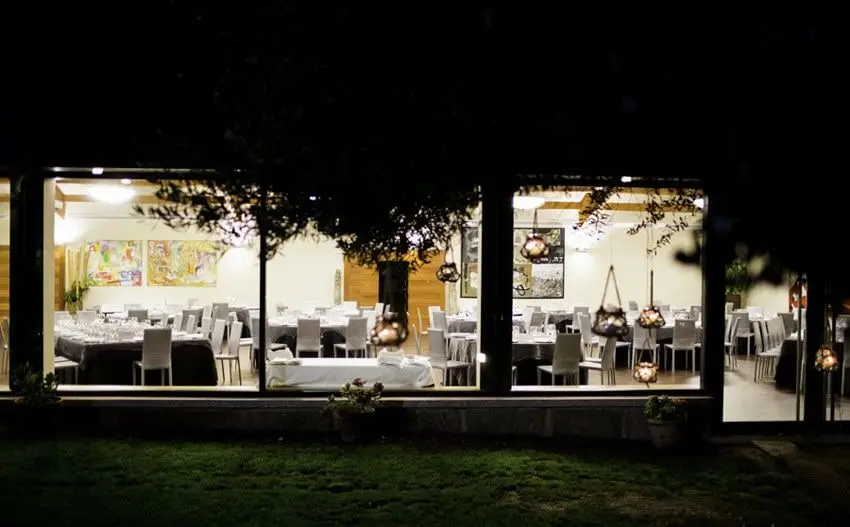 El salón de banquetes iluminado de noche visto a través de la cristalera, con las mesas puestas y un olivo delante