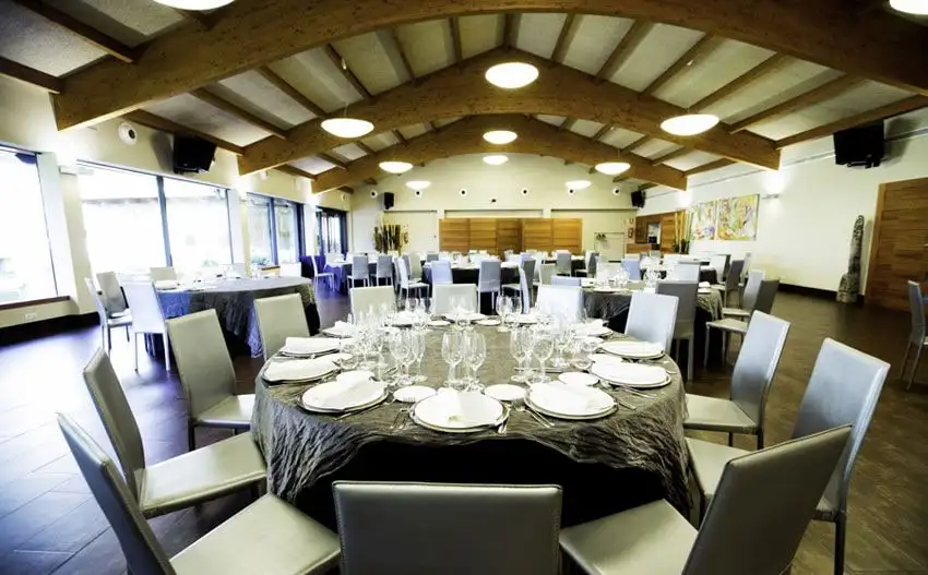 El salón de banquetes con vigas de madera vistas y una mesa redonda puesta, con cristalería y sillas grises, ante las cristaleras